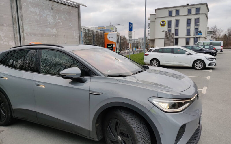 Graues Elektroauto an Ladesäule auf öffentlichen Parkplatz vor einem Hotel.